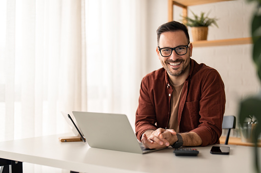 Smiling confident businessman looking at camera sitting at home office desk. Modern stylish corporate leader, successful CEO executive manager wearing business casual clothes and glasses posing for business portrait.