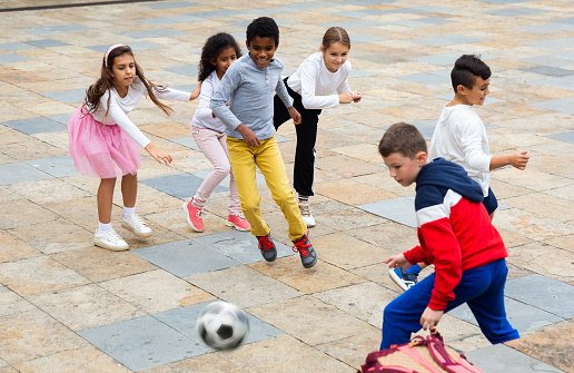 Cheerful tween schoolchildren gaily spending time together on warm spring day, playing with ball near school building