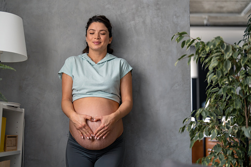 Dreamful expectant mother wearing soft blue t-shirt and gray leggings with the eyes closed holding her hands on belly and making a heart shape.