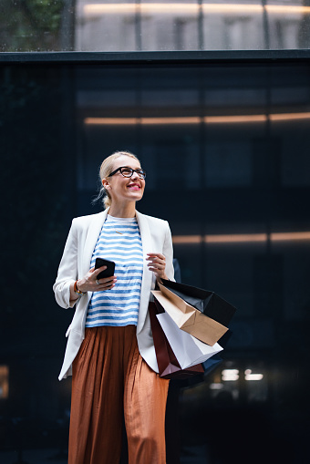 Smiling businesswoman typing text message on her smartphone and carrying paper bags while standing in the city.