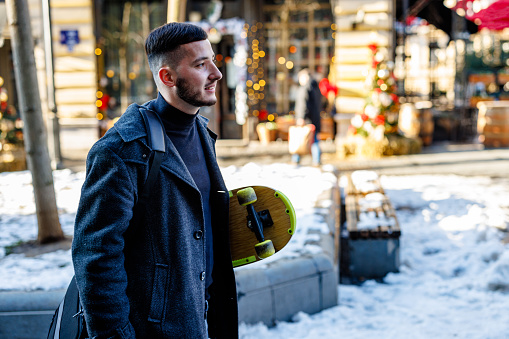 A young man is holding a skateboard and walking in the city district. A young handsome man is walking in the city street while carrying a skateboard on a cold winter day.