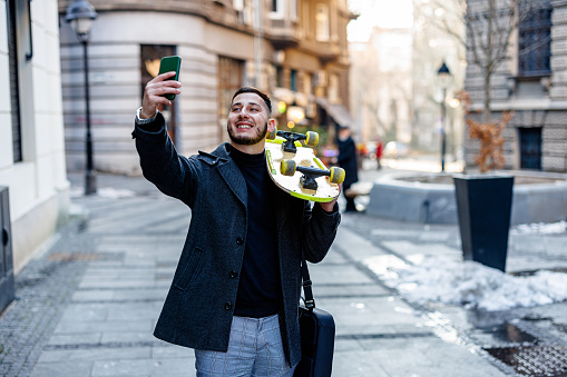 A happy smiling man in a business suit with a skateboard on the street holding a briefcase and using a mobile phone to take a self-portrait photo on a cold winter day.