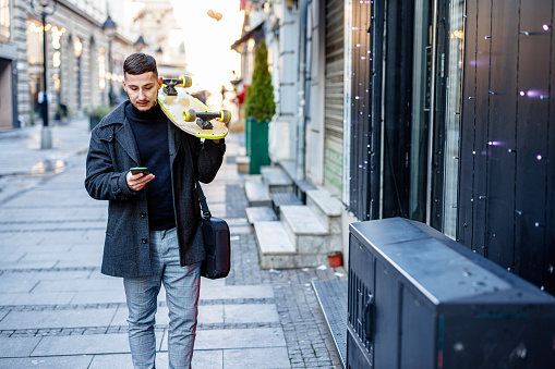 A happy smiling man in a business suit with a skateboard on the street holding a briefcase and using a mobile phone for text messaging on a cold winter day.