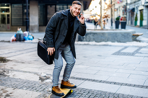 A happy smiling man in a business suit with a skateboard on the street holding a briefcase and using a mobile phone.