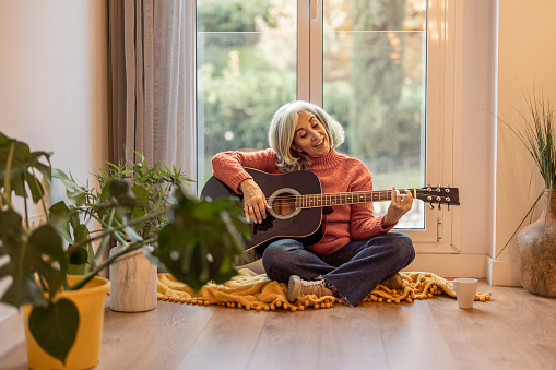 Senior woman playing guitar at home, grey-haired mature woman has fun at home by the window. life and home