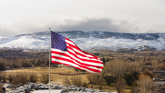 Aerial view of a Large American Flag in the Winter with rural farmland, snow capped mountains and a dramatic sky in the background.