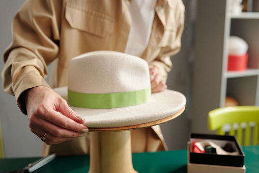 Close-up of hands of young craftswoman putting white felt hat with light green textile ribbon around its upper part on wooden equipment