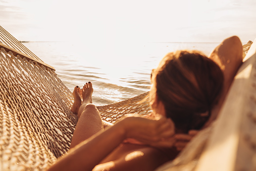 Young woman relaxing in wicker hammock on the sandy beach on Mauritius coast and enjoying sunset light over Indian ocean waves. Exotic countries vacation and mental health concept image.