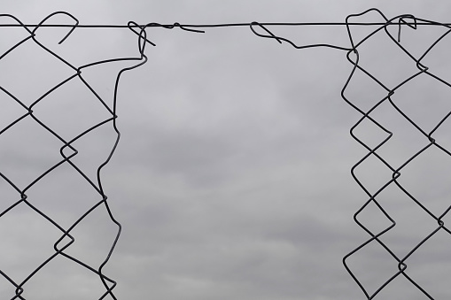 Chain link fence hole damage wire shapes and cloudy sky background.