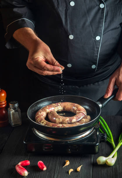 The chef adds salt to pan with raw meat sausage. Preparation for cooking sausages in the kitchen of a restaurant or cafe on table with vegetables and spices