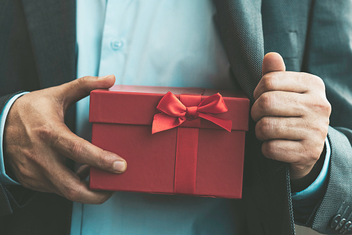 Chest view of an unrecognizable man wearing a suit who is about to give a red gift box out of his jacket. The red gift box is wrapped with red ribbon.  Mother’s day / Valentine’s day / Promotion concept.