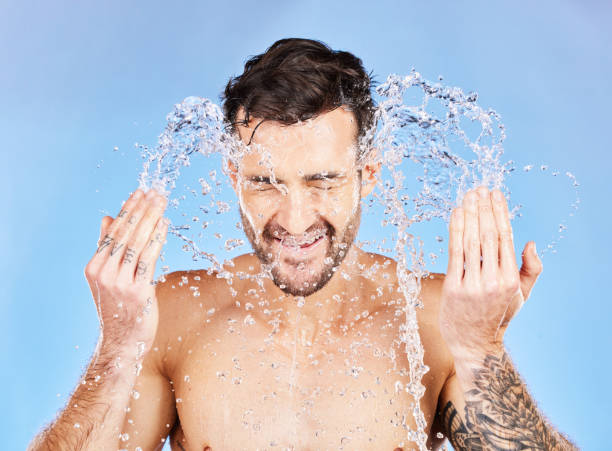 istock Face, water and cleaning with a man model splash in studio on a blue background for hygiene. Skincare, water splash and hydrate with a handsome young male washing his skin in the bathroom for care 1456635802