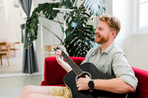 Ginger man playing guitar and sitting on red sofa at home
