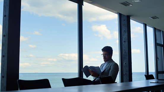 Lawyer studying contract papers at sea panorama window. Focused man reading deal documents in conference hall. Confident businessman executive check financial annual report. Male professional concept