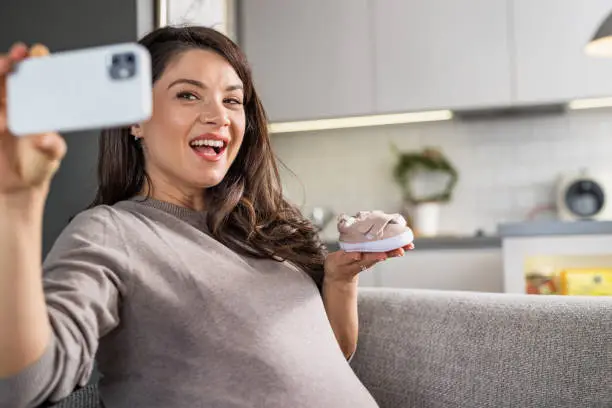 Lovely pregnant brunette holding baby booties and taking a selfie with her smartphone. Expecting a new life concept
