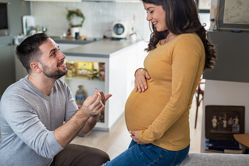 A man with an engagement ring is kneeling and proposing marriage to a pregnant woman