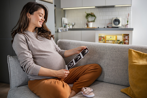 Portrait of a pregnant woman, who is about to give birth, looking at an ultrasound scan. She is sitting on the couch in the lotus position. Baby booties are on the couch.