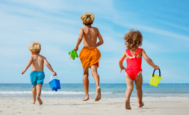 trois enfants heureux courent sur la plage de sable avec des seaux par derrière - shorts rear view summer beach photos et images de collection