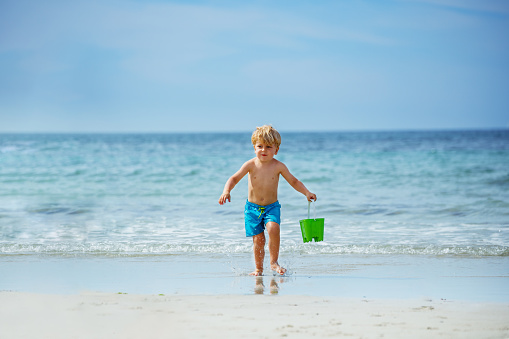 Little blond boy run to ocean waves on sand beach carry toy bucket with water