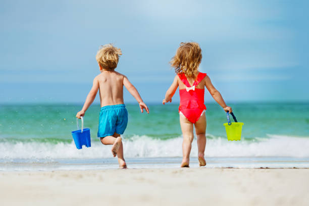 vue de derrière d’un garçon et d’une fille qui courent avec un seau jusqu’à l’océan - shorts rear view summer beach photos et images de collection