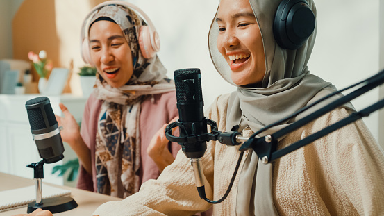 Happy muslim Asian girl friends wear hijab and headphone record podcast on laptop talk microphone onair live stream with audience and guest at living room. Female podcaster audio podcast home studio.