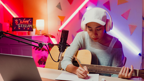 Happy Asian girl influencer wear headphone record podcast on laptop play guitar writing notes to music book onair online live streaming at studio room. Female podcaster audio podcast home studio.