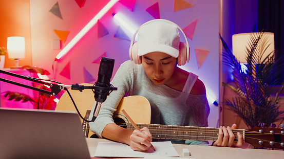 Happy Asian girl influencer wear headphone record podcast on laptop play guitar writing notes to music book onair online live streaming at studio room. Female podcaster audio podcast home studio.