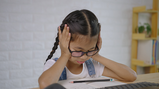 Children who are bored with learning online with a laptop at home, an Asian girl 9 years old with a hand on her head, Studying online, Home School