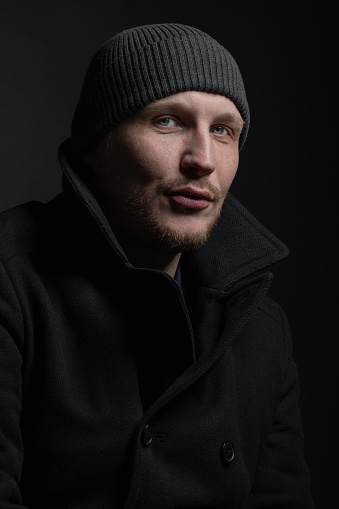 Lifestyle, fashion, occupation concept. Man with winter hat and black coat studio portrait. Model with beard looking at camera with serious look. Dark studio background