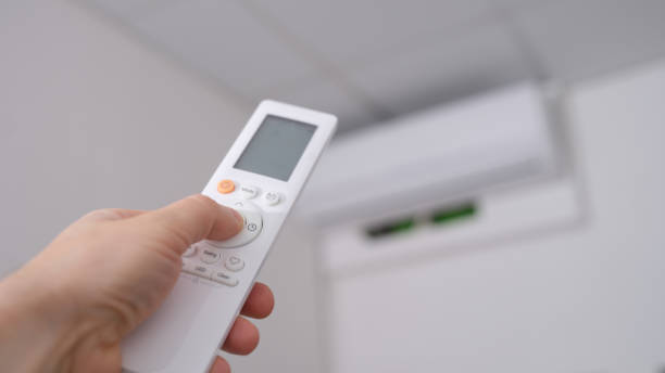 la main d’une femme tient une télécommande pointant vers le climatiseur dans la pièce - air conditioner photos et images de collection