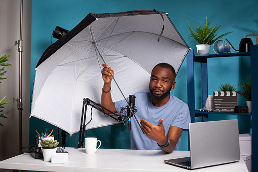 Technology reviewer testing flash reflector umbrella presenting size and benefits for live vlog broadcast. Professional photographer vlogger reviewing photography studio equipment in live podcast.
