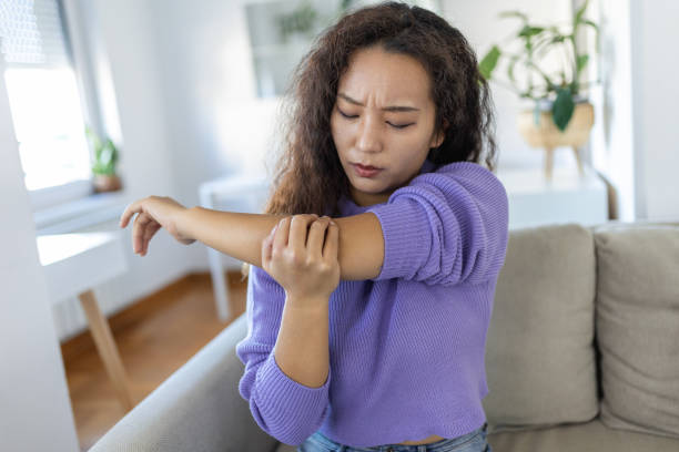 unglückliche asiatische frau mit ellenbogenschmerzen in innenräumen. bürosyndrom gesundheitskonzept. verärgert stirnrunzelnde asiatische dame verwirrt beim betrachten der schmerzen auf der couch zu hause in gemütlicher heller wohnung. - ellenbogen stock-fotos und bilder