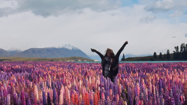 Young happy woman dancing in field of bright colourful lupin flowers with blue lake and mountain background