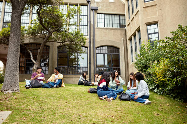 Young friends taking a break between college classes Men and women in teens and 20s sitting on grass, talking, eating lunch, and checking smart phones. Property release attached. campus stock pictures, royalty-free photos & images