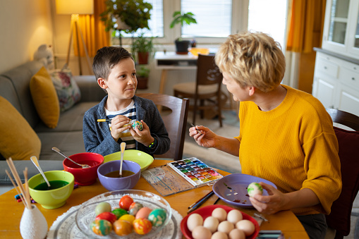 A mother is looking at her son while they are painting Easter eggs at the table, with eggs ready to paint and bowls of dye to color the eggs