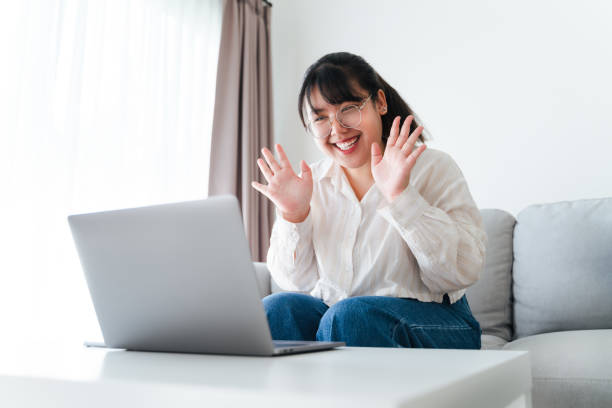 istock Young Asian woman using laptop computer for online video conference call waving hand making hello gesture on the couch in living room. 1456734435