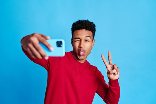 Cheerful African American male in red shirt taking self portrait on smartphone against blue background
