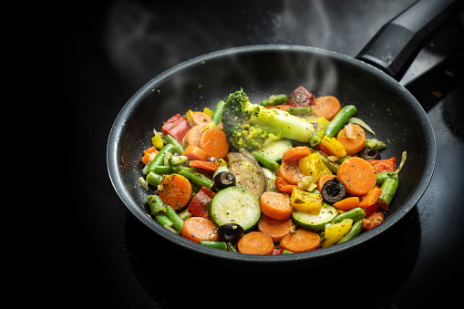 Stir fry vegetables in a black pan with carrots, zucchini, broccoli, green beans, olives and herbs, healthy vegetarian meal Mediterranean style, copy space, selected focus, narrow depth of field