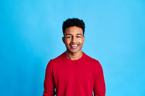 Positive African American male in red shirt smiling and looking at camera while standing against blue background