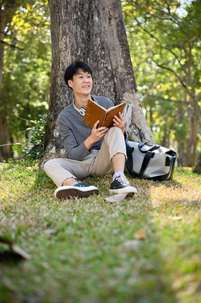 istock Handsome Asian male college student reading a book under the tree in greenery park. 1456741323