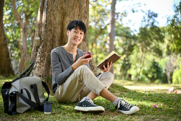 istock Asian male college student sits under the tree in the park, eating apple while reading a book 1456741385