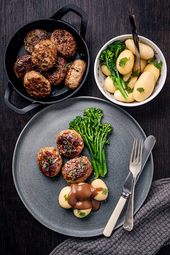 Recipe with pan fried  Danish meat balls or frikadeller in Danish. Served with boiled new potatoes and sprouting broccoli spears. They are a staple of the Danish diet and they are traditionally made from a mix of veal and pork, herbs and spices such as sage and thyme as well as salt and pepper. Colour, overhead view vertical with some copy space.