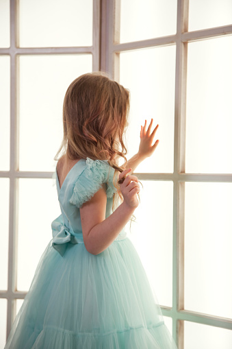 Rear view sad little adorable girl in art azure dress posing near large window indoors, showing excitement. Studio shot unhappy pensive kid lady. Child emotion concept. Copy text space for advertising