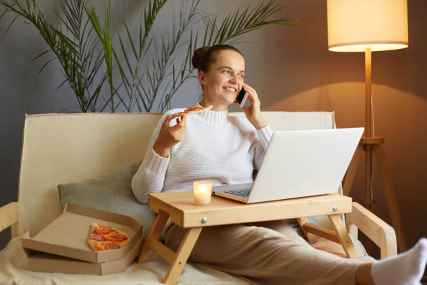 на снимке в помещении женщина расслабляется, проводя выходной день дома, используя блокнот, поедая пиццу и разговаривая по мобильному теле� - проводя поиск стоковые фото и изображения