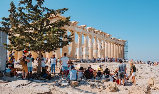 Athens, Greece - August 30, 2022: Visitors protecting themselves from the intense sun of a summer day in the shade of a tree, in front of the famous Parthenon, in the Acropolis of Athens, Greece
