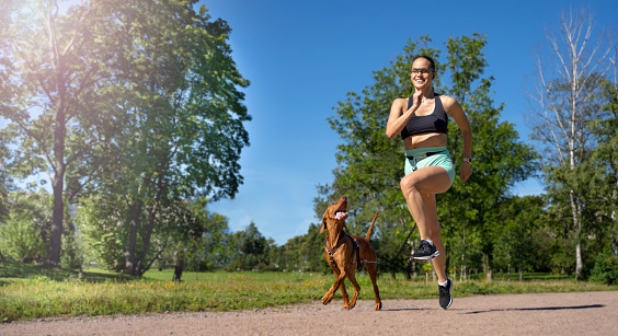 Sporty happy woman jogging with dog outdoor in public park summer. Canicross competition. Healthy life with active pet. Copy space for design