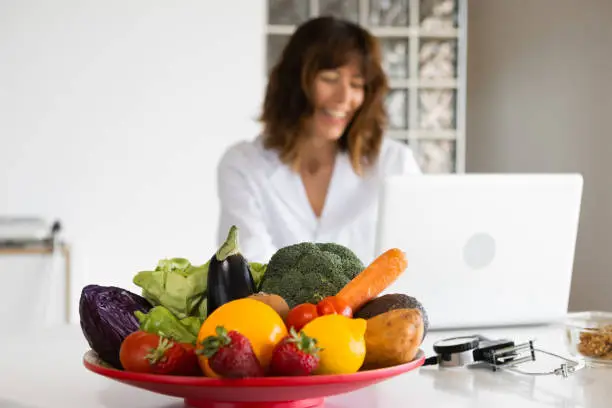 Female nutritionist working with laptop in a doctor's office  with big fresh vegetable and fruit plate. Concept of a healthy lifestyle, body care and weight loss. Selective focus. Blurred background.