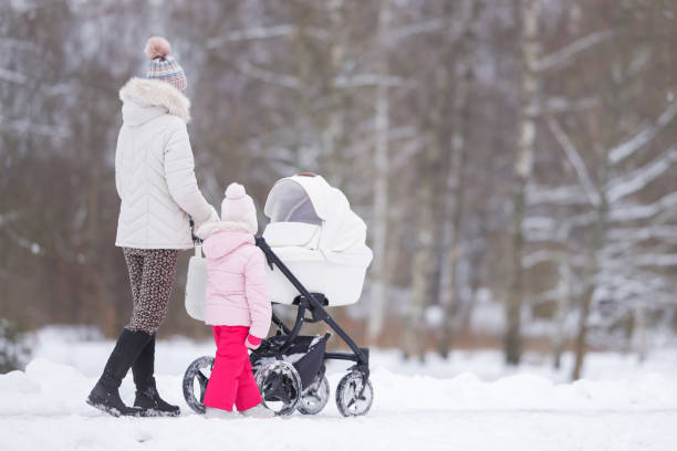 junge erwachsene mutter und kleine tochter schieben weißen kinderwagen und gehen an kalten wintertagen auf dem schneebedeckten bürgersteig im park. zeit miteinander verbringen. genießen sie den spaziergang. zwei kinder mutter. rückansicht. - snow park road cold stock-fotos und bilder