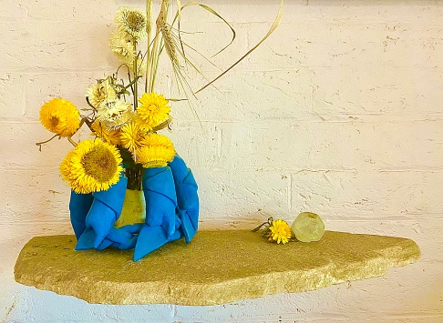 Horizontal still life of material vase with dried yellow daisy wild flowers picked from headland grass, on wood wall bench against painted brick wall in country Mullumbimby NSW Australia