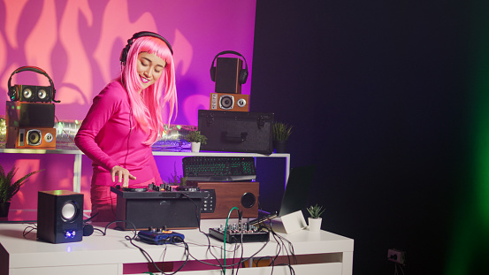 Asian musician with headphones playing electronic song at mixer console, standing at dj table having fun with fans at night in club. Artist performing techno music in studio over pink background